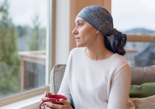 A young woman wearing a headscarf sits in a living room, holding a ceramic mug while looking out the window.