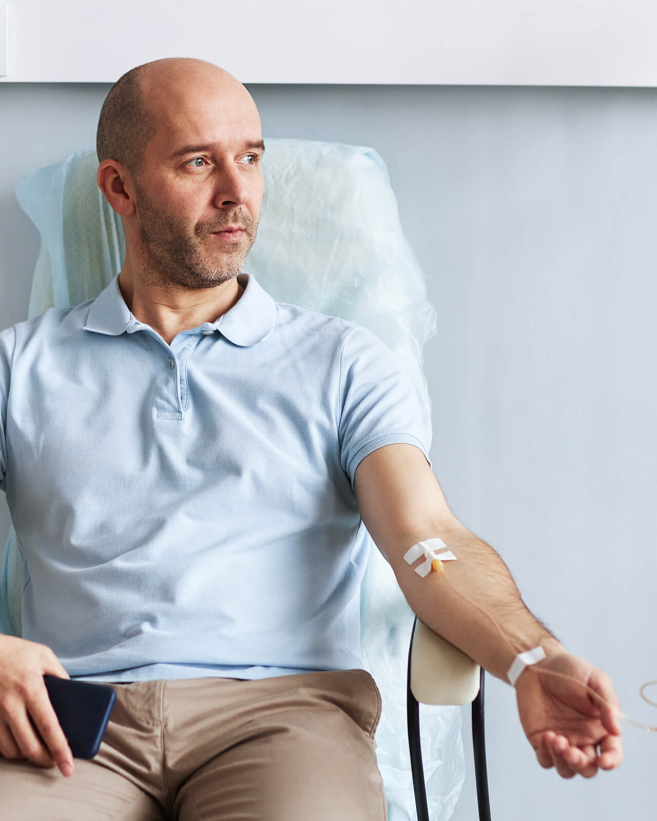 A man sitting in a chair receiving chemotherapy, gazing off to the right with a mobile device in his hand.