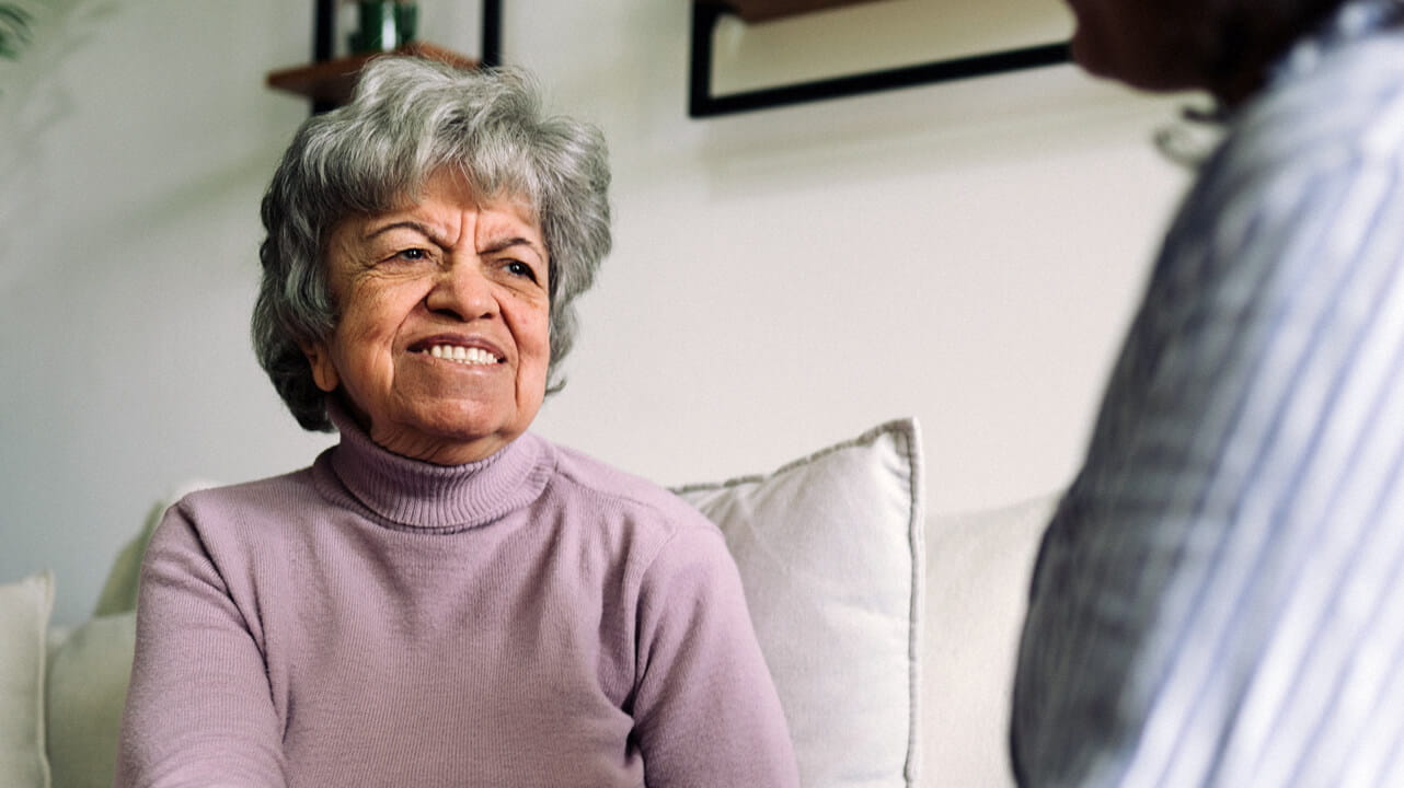 An older woman sitting on a couch, holding hands with someone off camera.