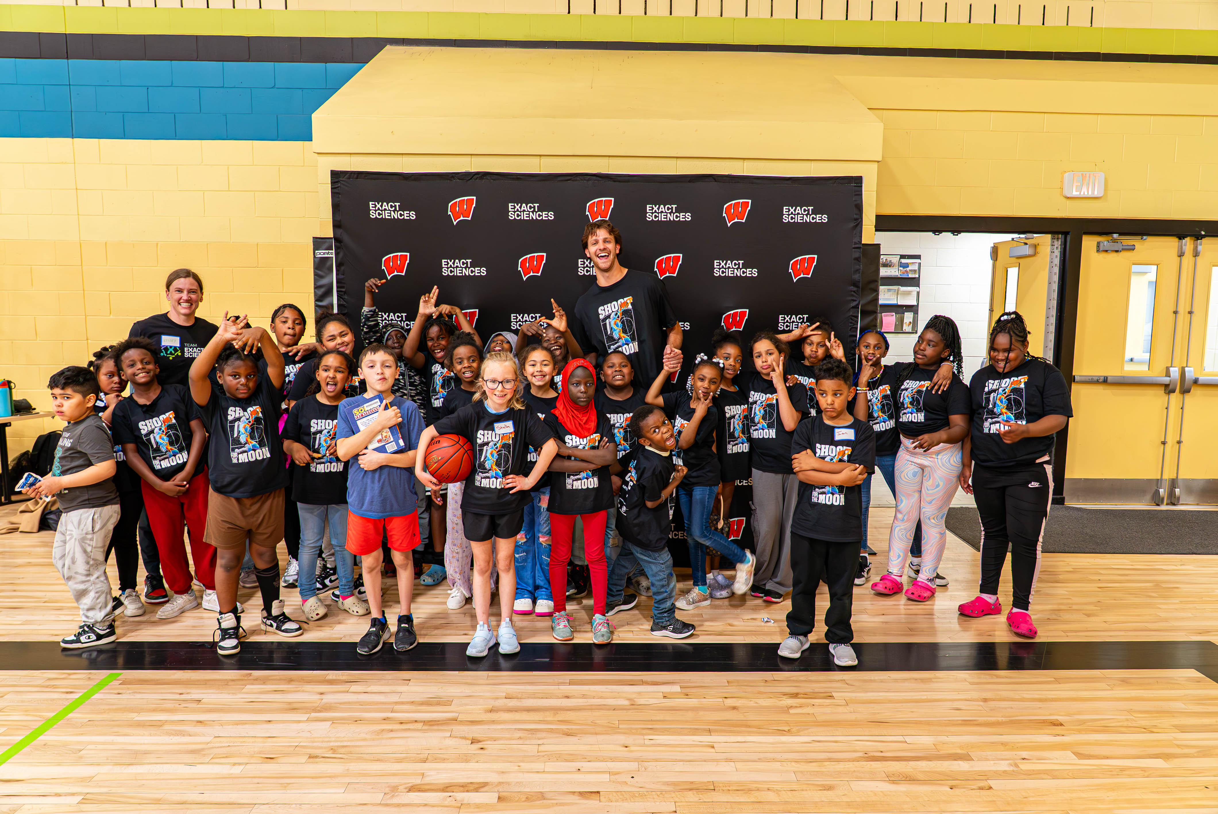 A group of children smiling and posing together for a photo in a gym setting with University of Wisconsin Badger basketball star Carter Gilmore and Exact Sciences staff